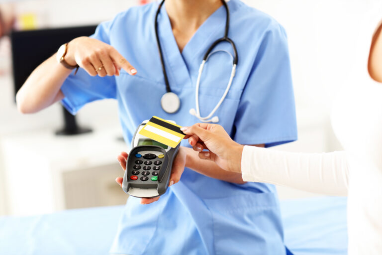 Allied health professional in blue scrubs holding an EFTPOS terminal while a patient taps a card to pay at a clinic