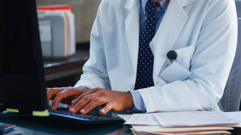 Doctor in a white coat typing on a computer keyboard at a desk with patient files, managing online appointments through a booking platform such as HotDoc or HealthEngine